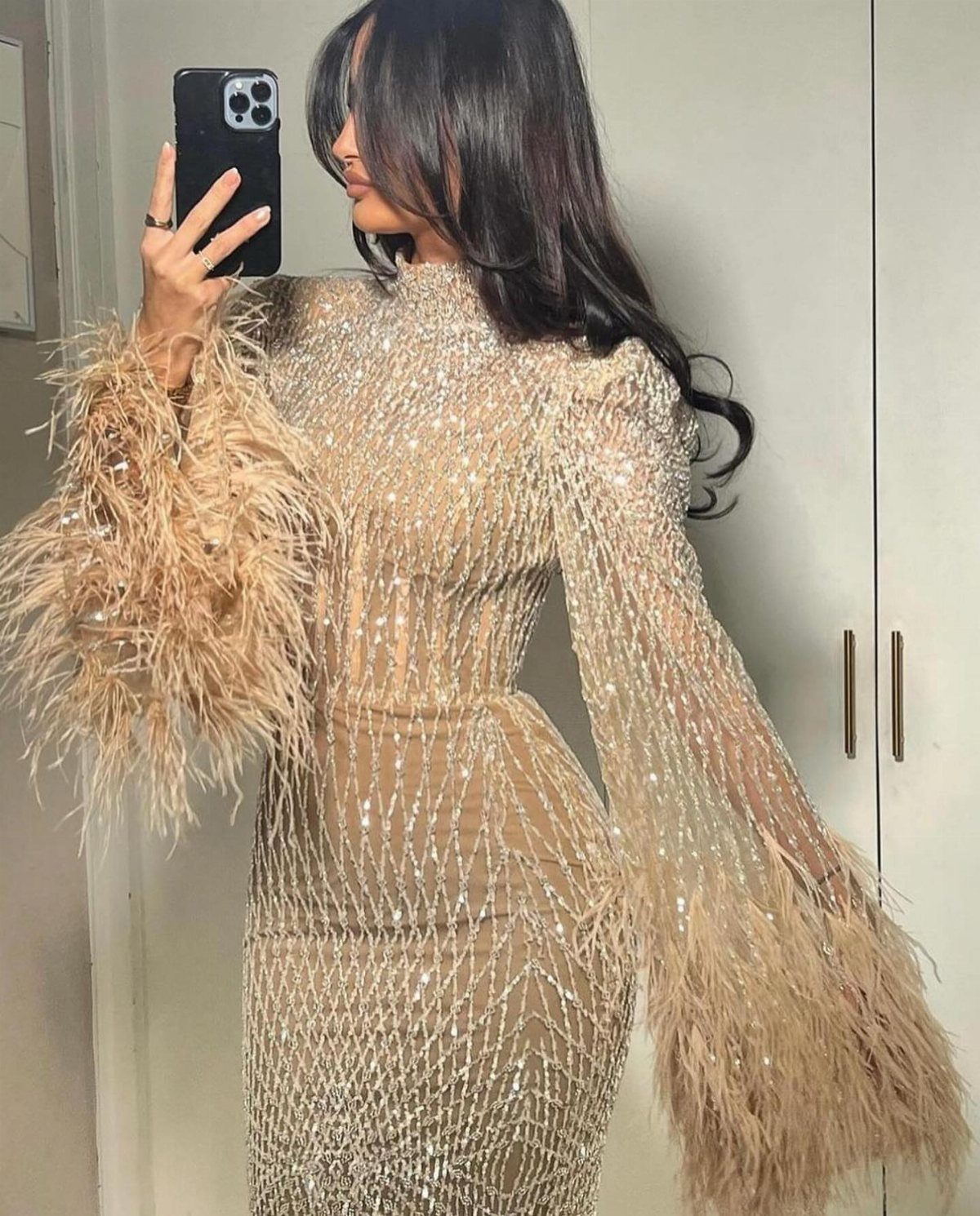 Woman in a sequined champagne long sleeve dress with feather cuffs taking a selfie.