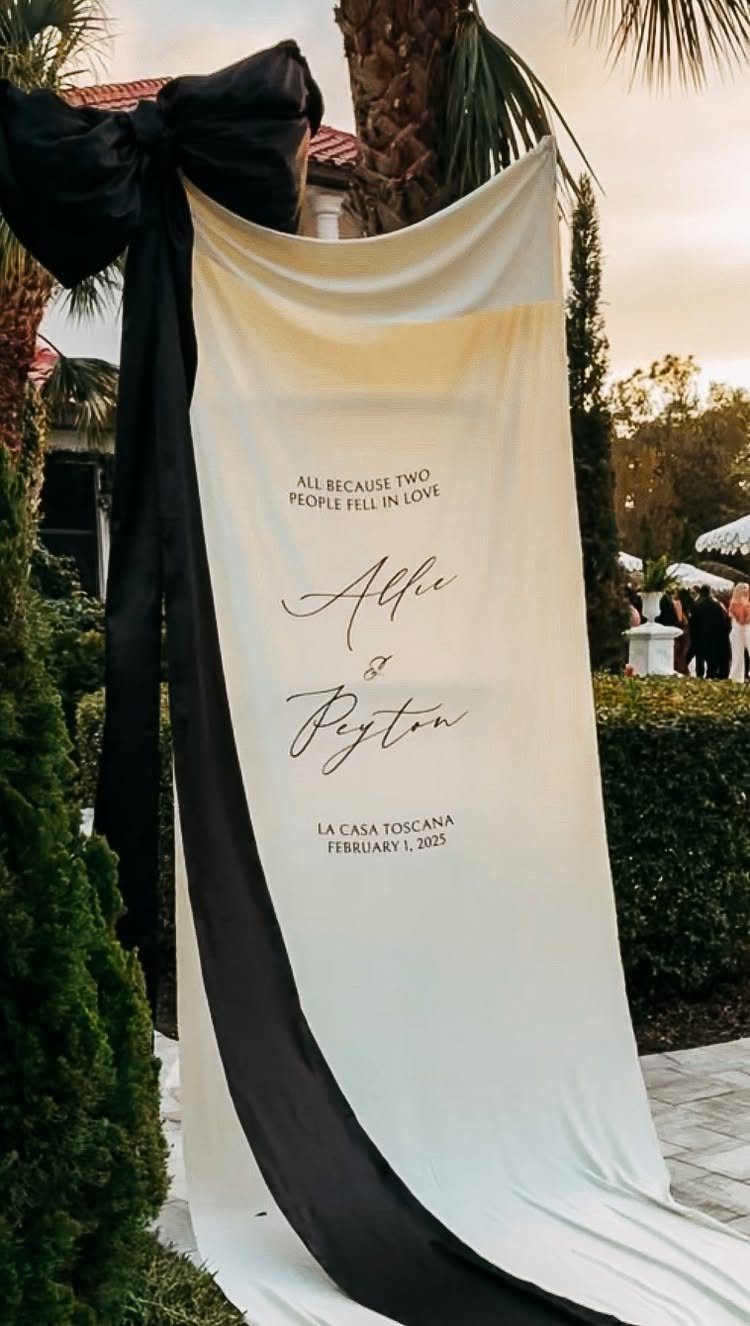 White fabric wedding banner with black ribbon and elegant calligraphy draped outdoors.