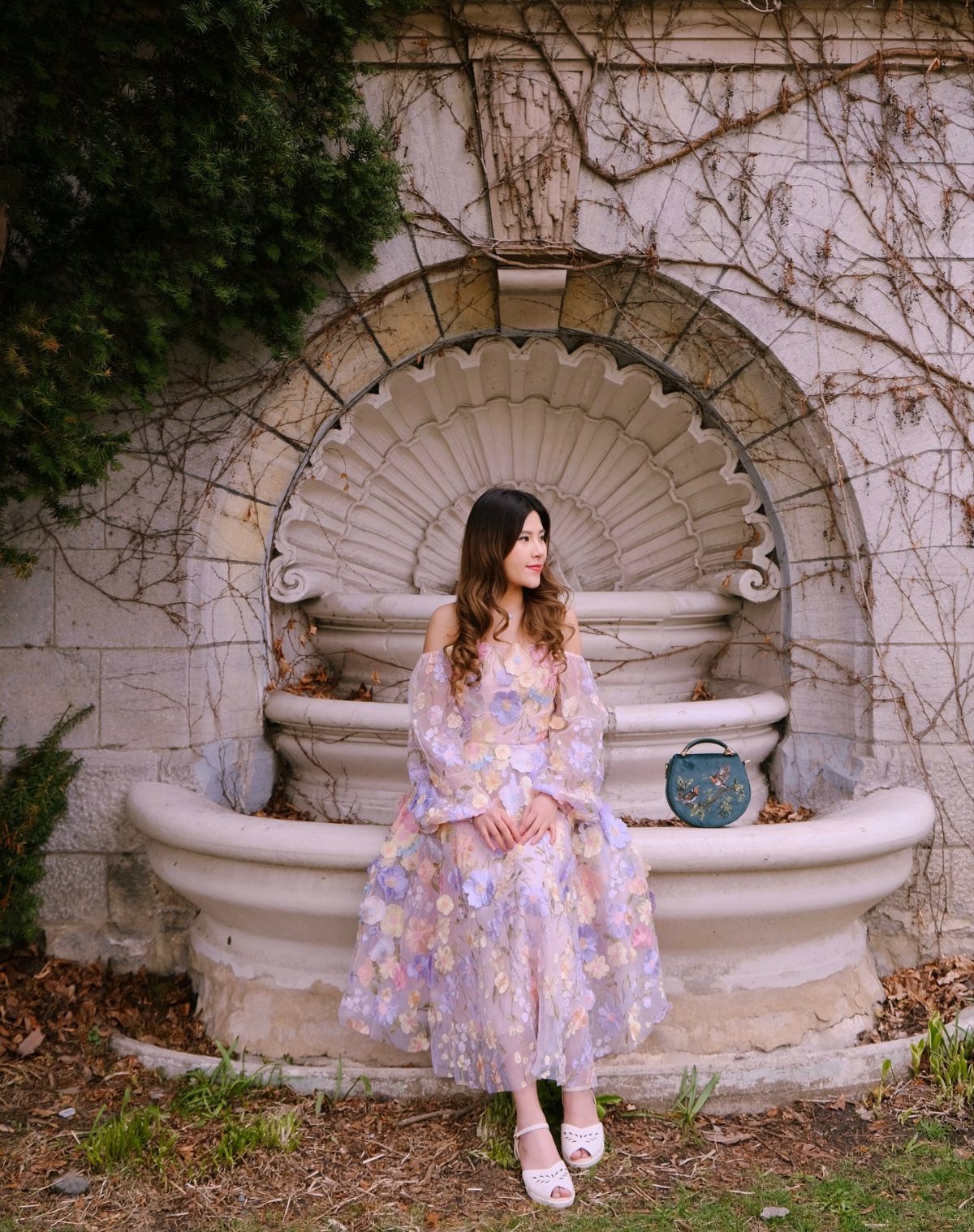 Woman in a purple floral midi dress sitting on a stone shell fountain.
