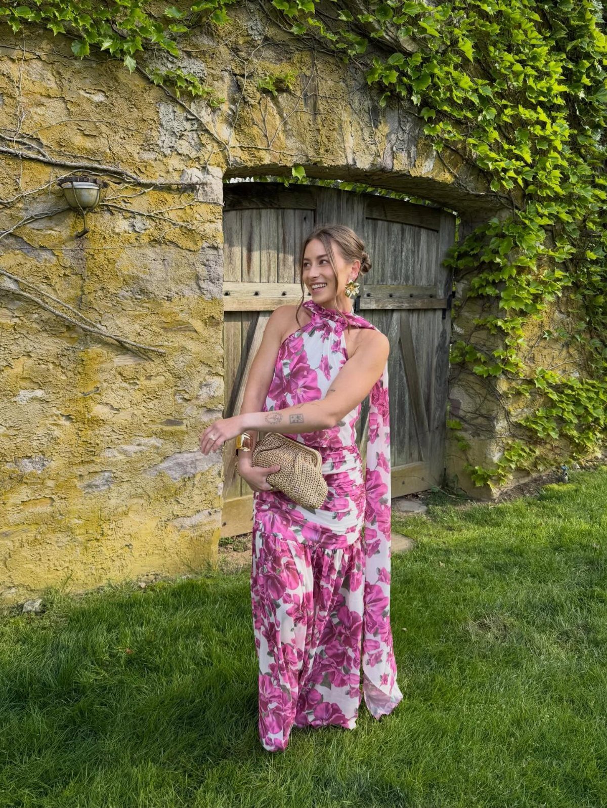 Woman in a pink floral one-shoulder maxi dress holding a straw bag outdoors.