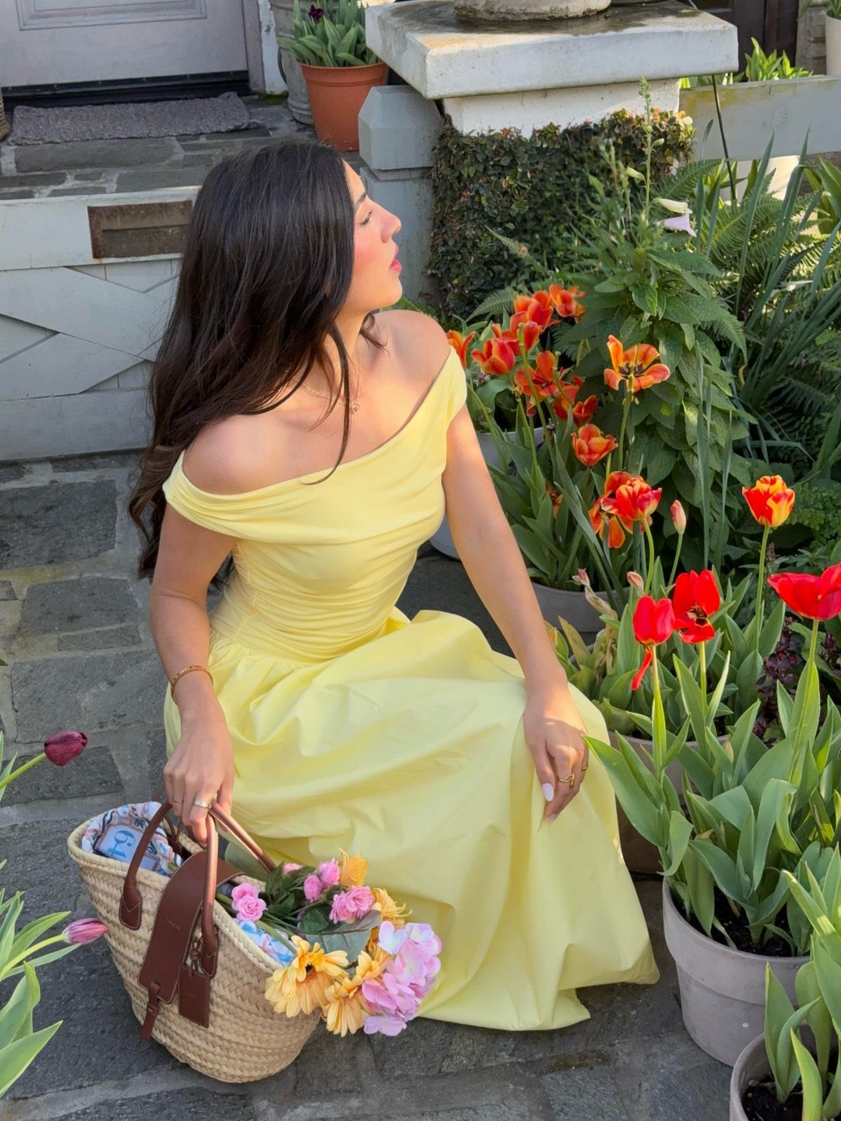 Woman in a yellow off-the-shoulder dress sitting by red tulips with a straw bag.