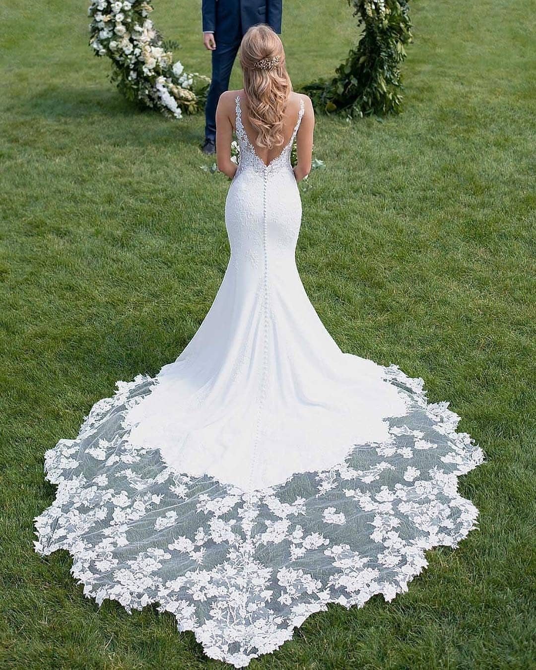 Woman in a white mermaid-style wedding dress with a long lace train on grass.