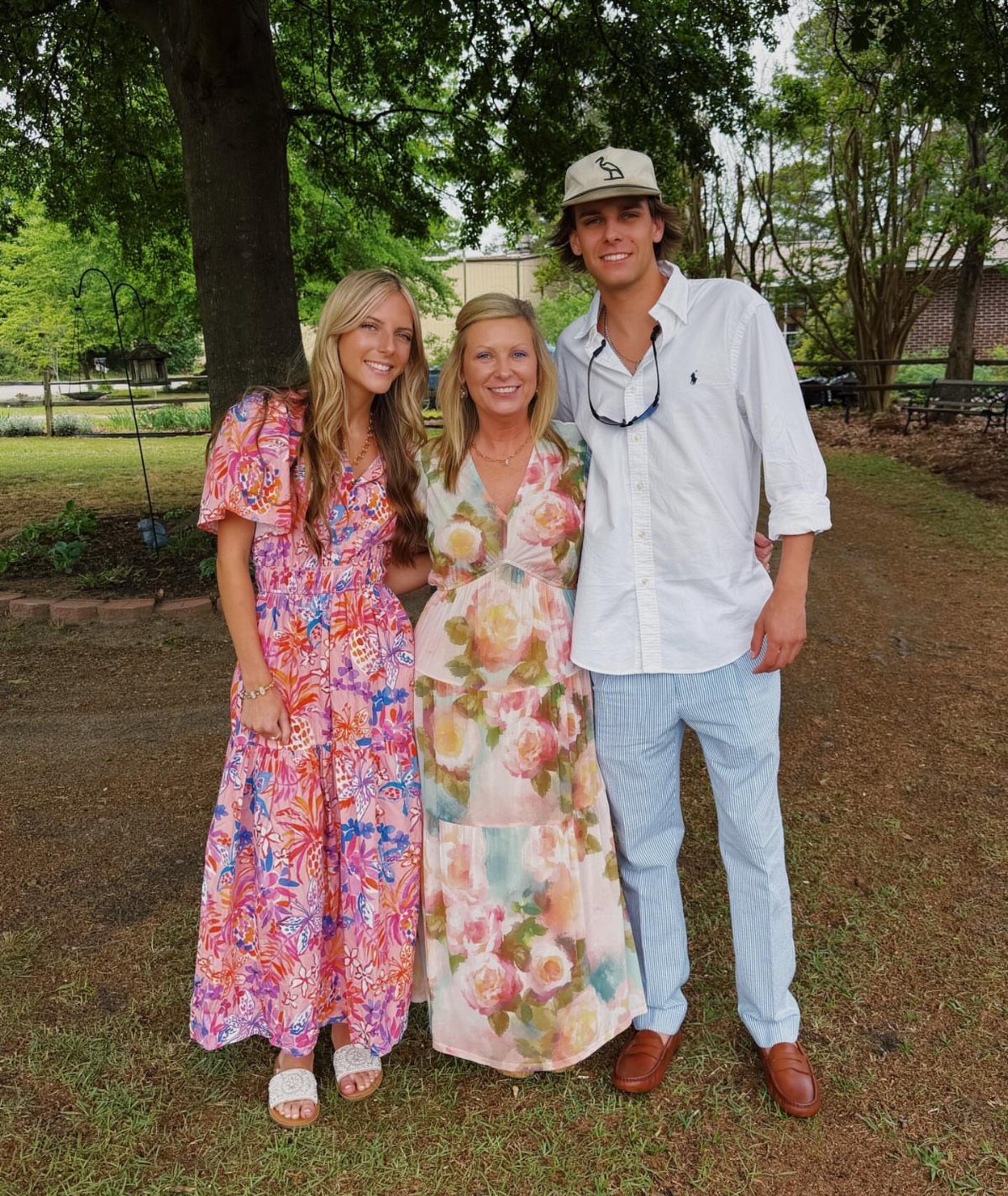 Three people wearing floral dresses and a white shirt with striped pants outdoors.