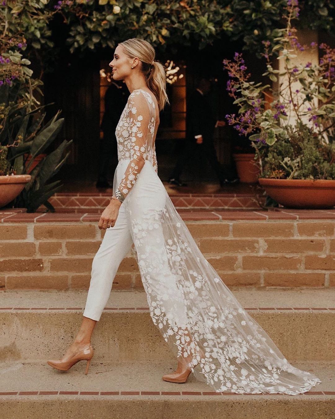 Woman in a white lace bridal jumpsuit with a long train on brick steps.