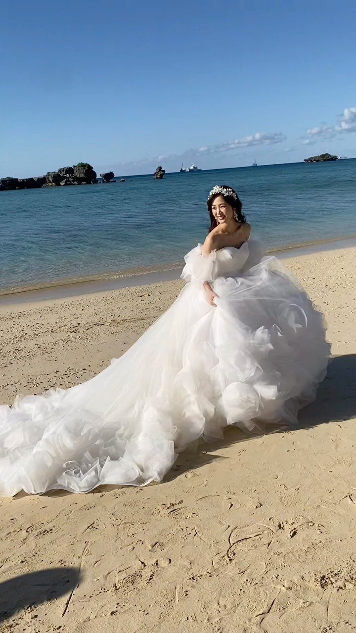 Woman in a voluminous white wedding gown and floral headpiece on a sandy beach.