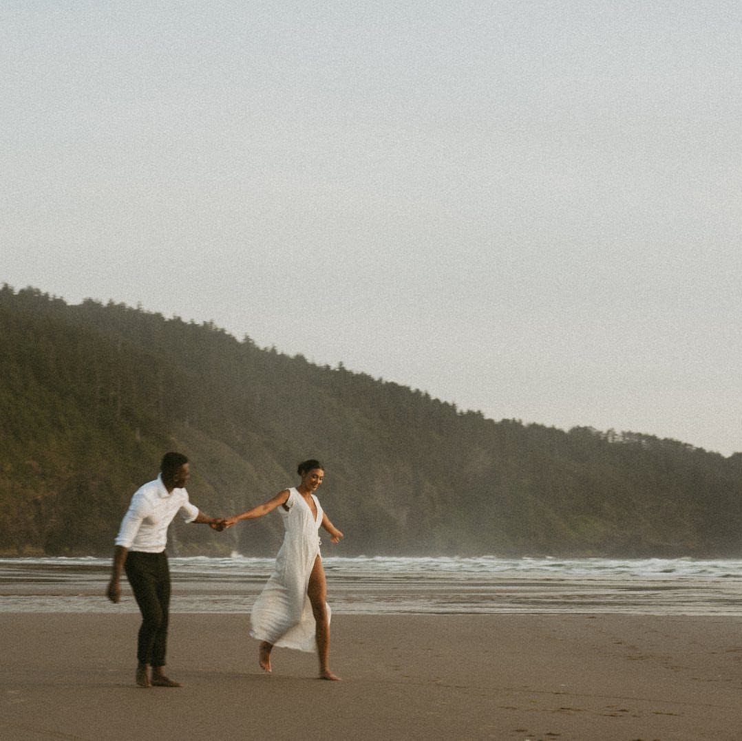 Couple in white beach attire holding hands and walking on a sandy shore.