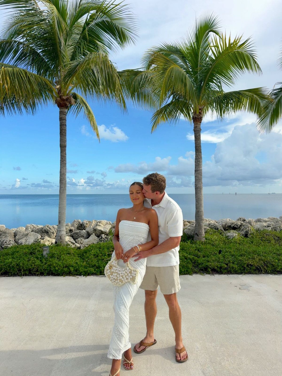 Couple posing by the ocean, woman in a white strapless sundress and man in beige shorts.