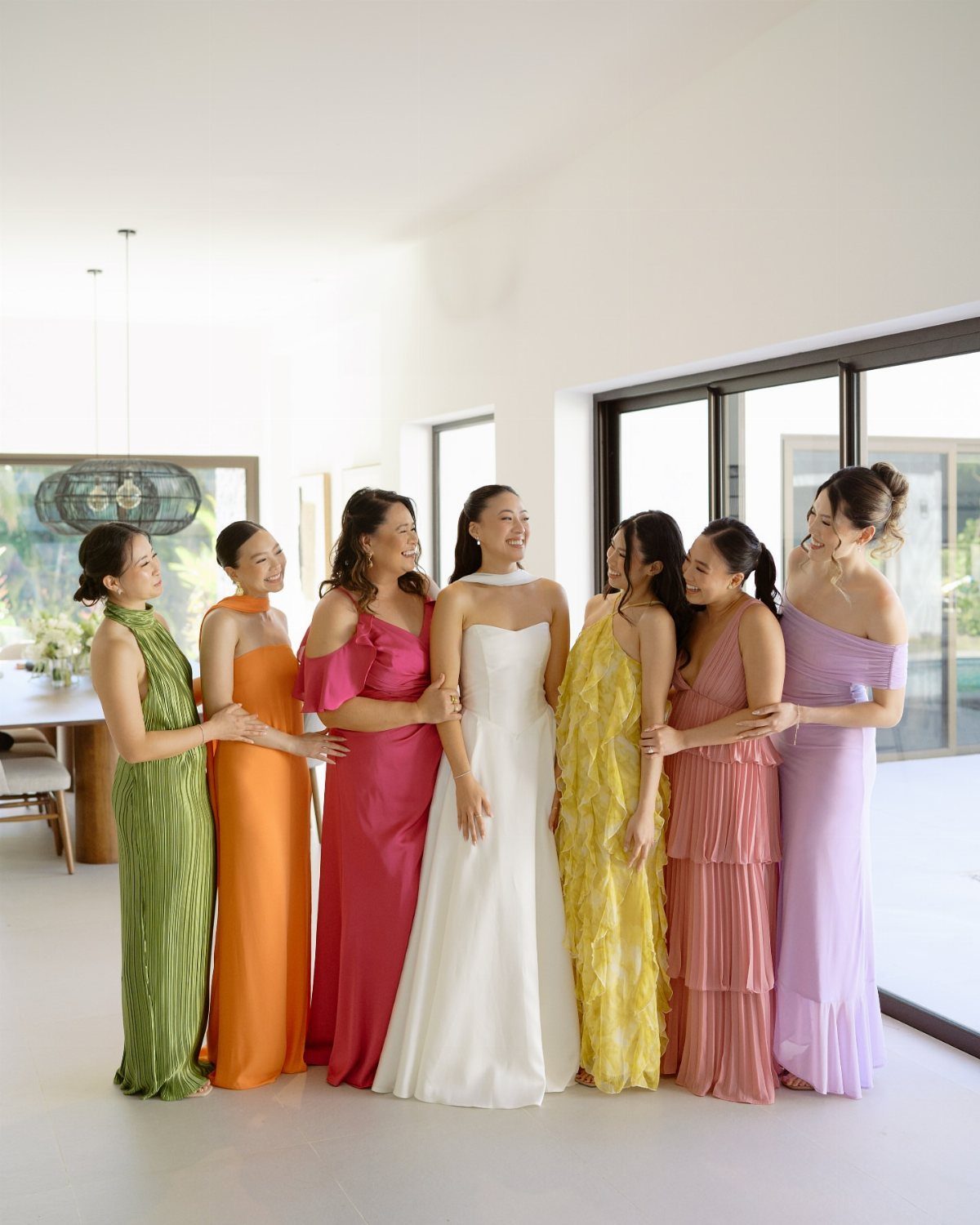 Seven women in colorful long gowns posing together in a bright modern room.