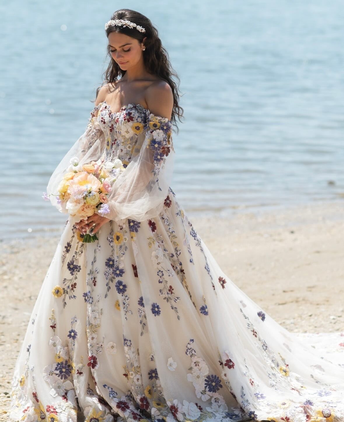 Woman in a white floral embroidered wedding dress holding a bouquet on a beach