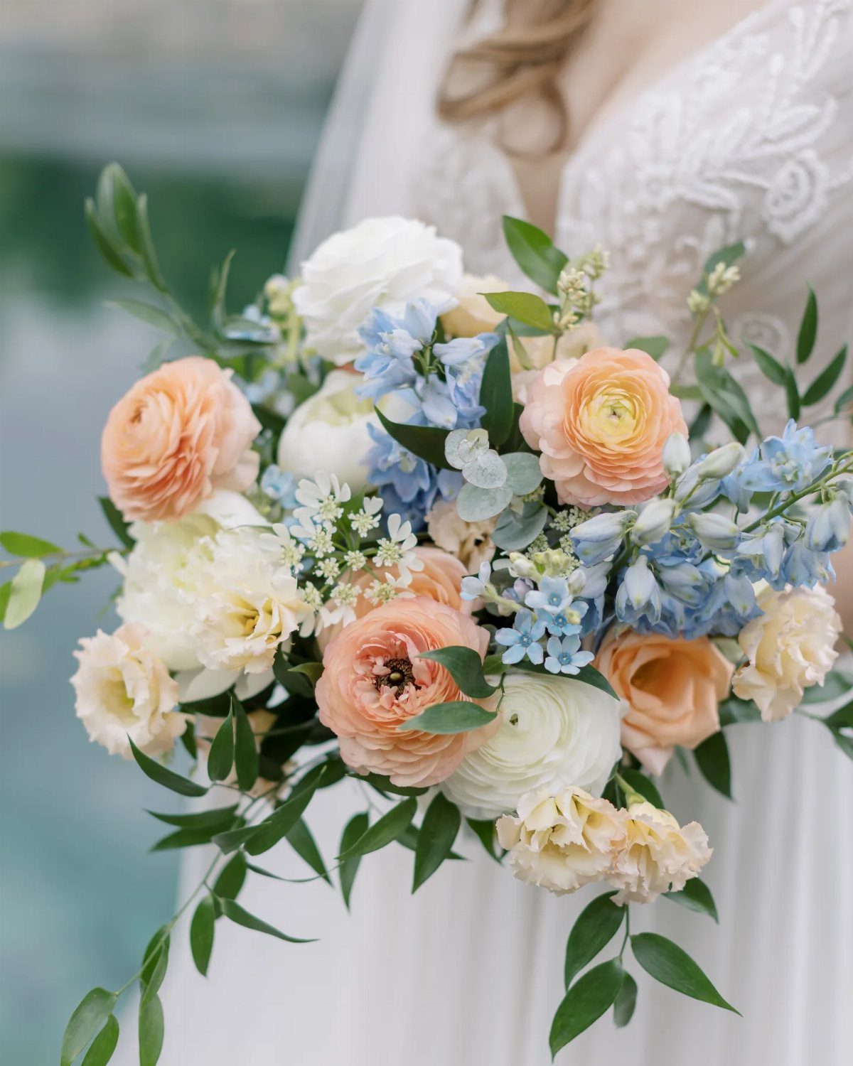 Bridal bouquet with peach, white, and light blue flowers against a white lace dress.