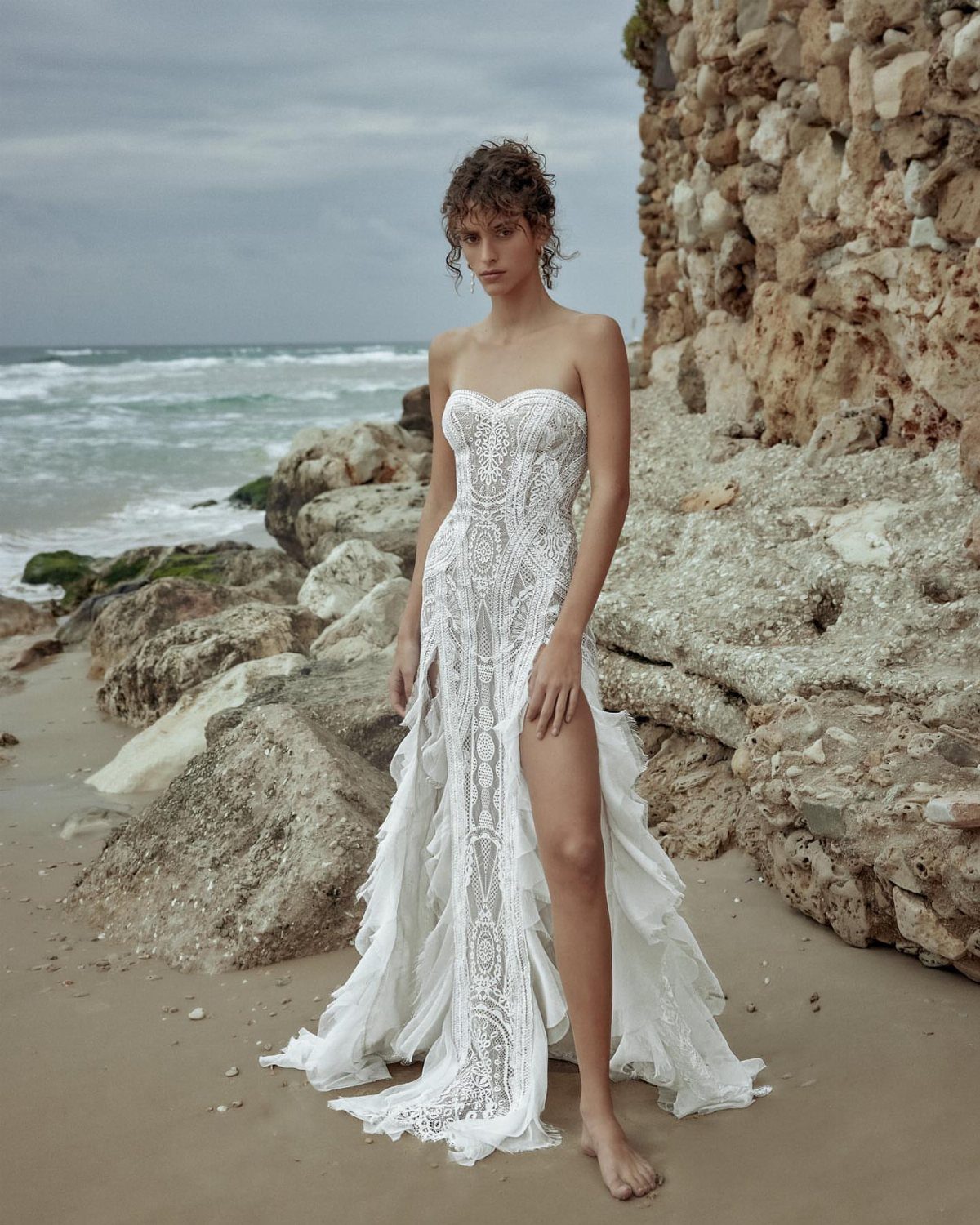 Woman in a white bohemian lace wedding dress standing on a rocky beach.