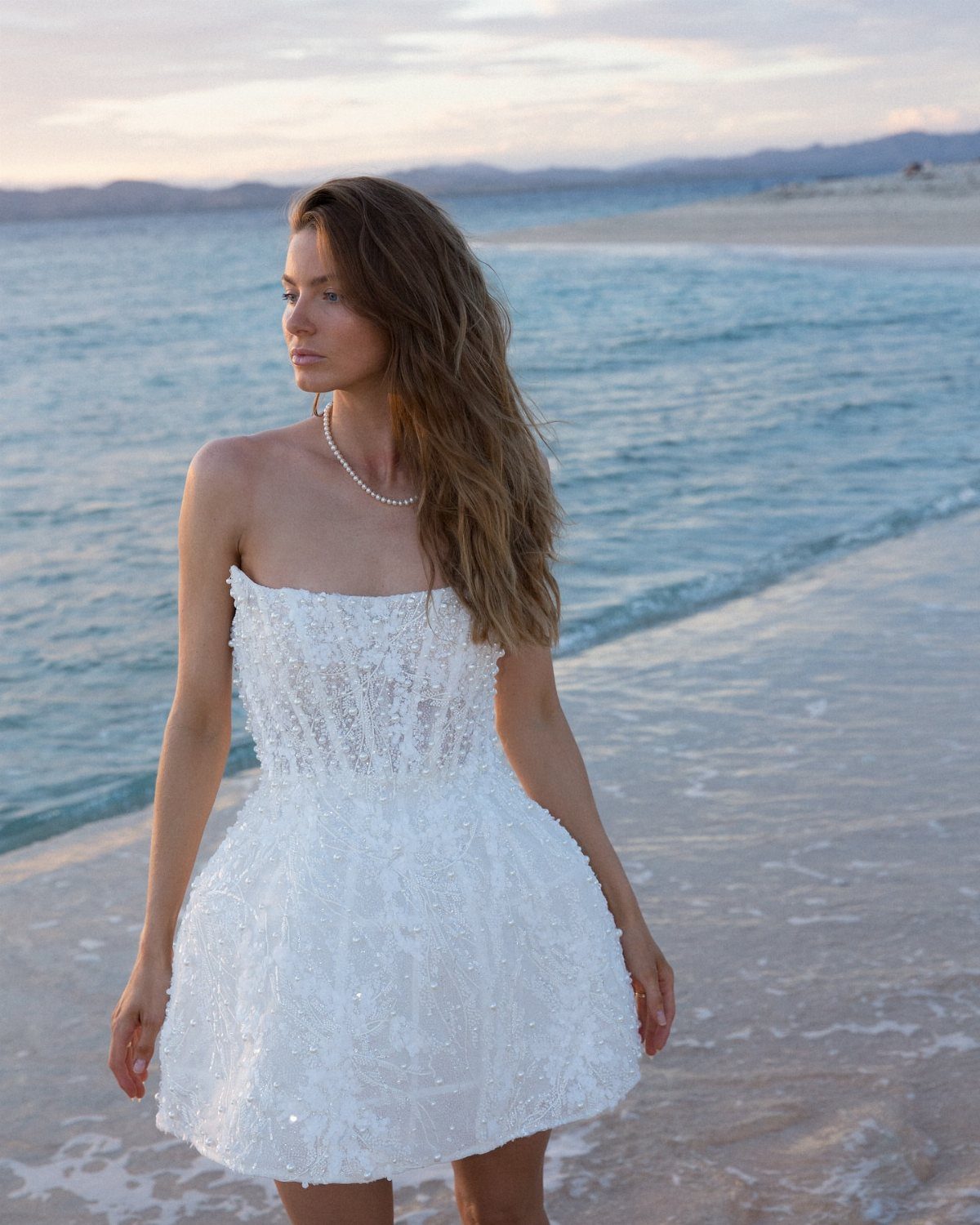 Woman in a short strapless white sequined wedding dress standing on a sandy beach.