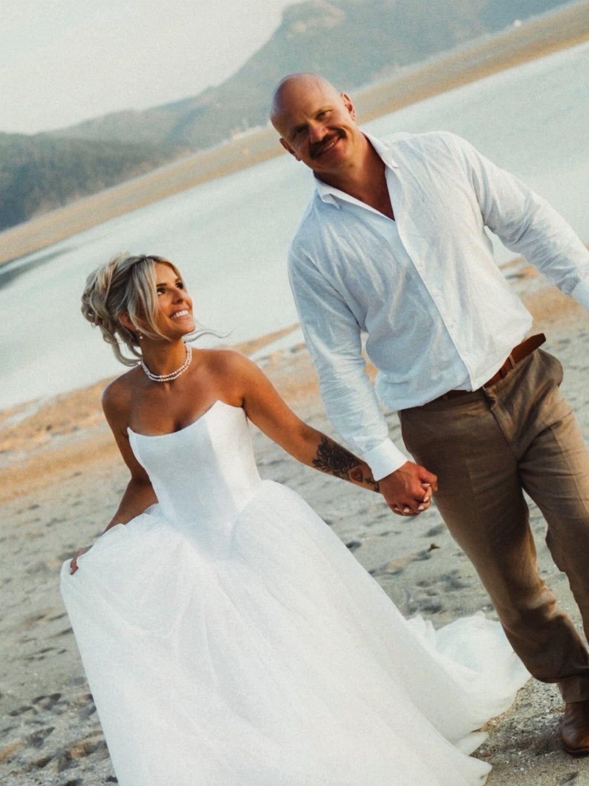 Bride in a strapless white wedding gown and groom holding hands on a beach.