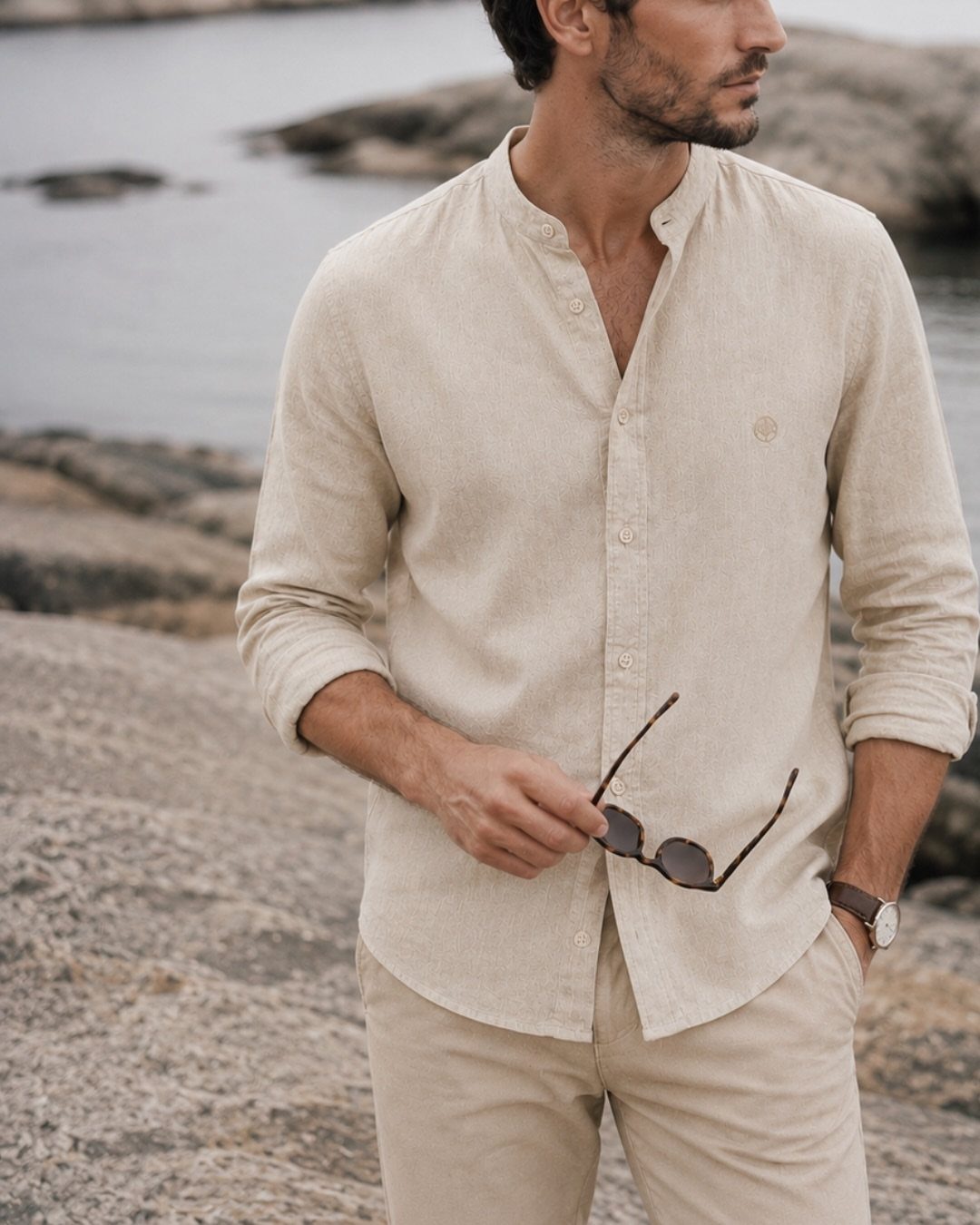 Man in a beige linen shirt and cream trousers standing on a rocky beach.