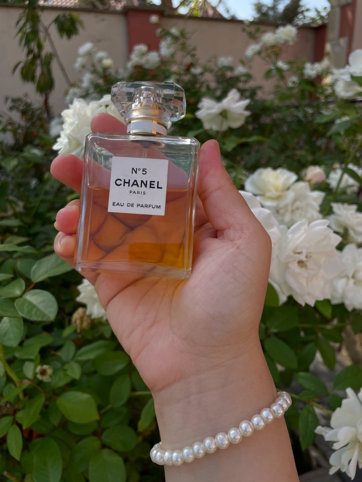 Chanel No. 5 perfume bottle held by a hand against white roses.