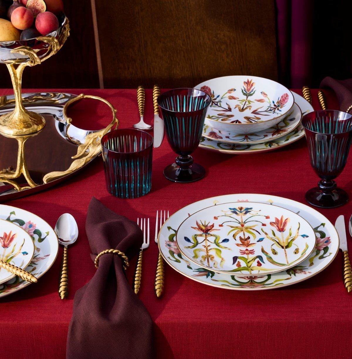 Floral patterned white dinnerware and blue glassware on a red tablecloth with gold cutlery.