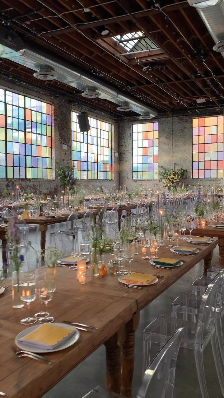 Wooden wedding tables with acrylic chairs in an industrial room with colorful windows.