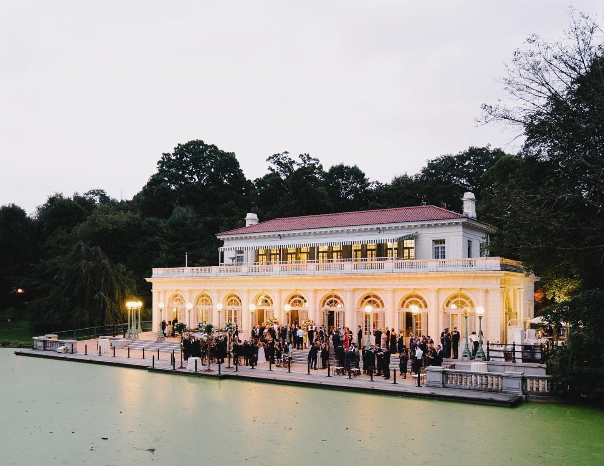 Historic white boathouse venue with guests on a terrace beside a green pond.