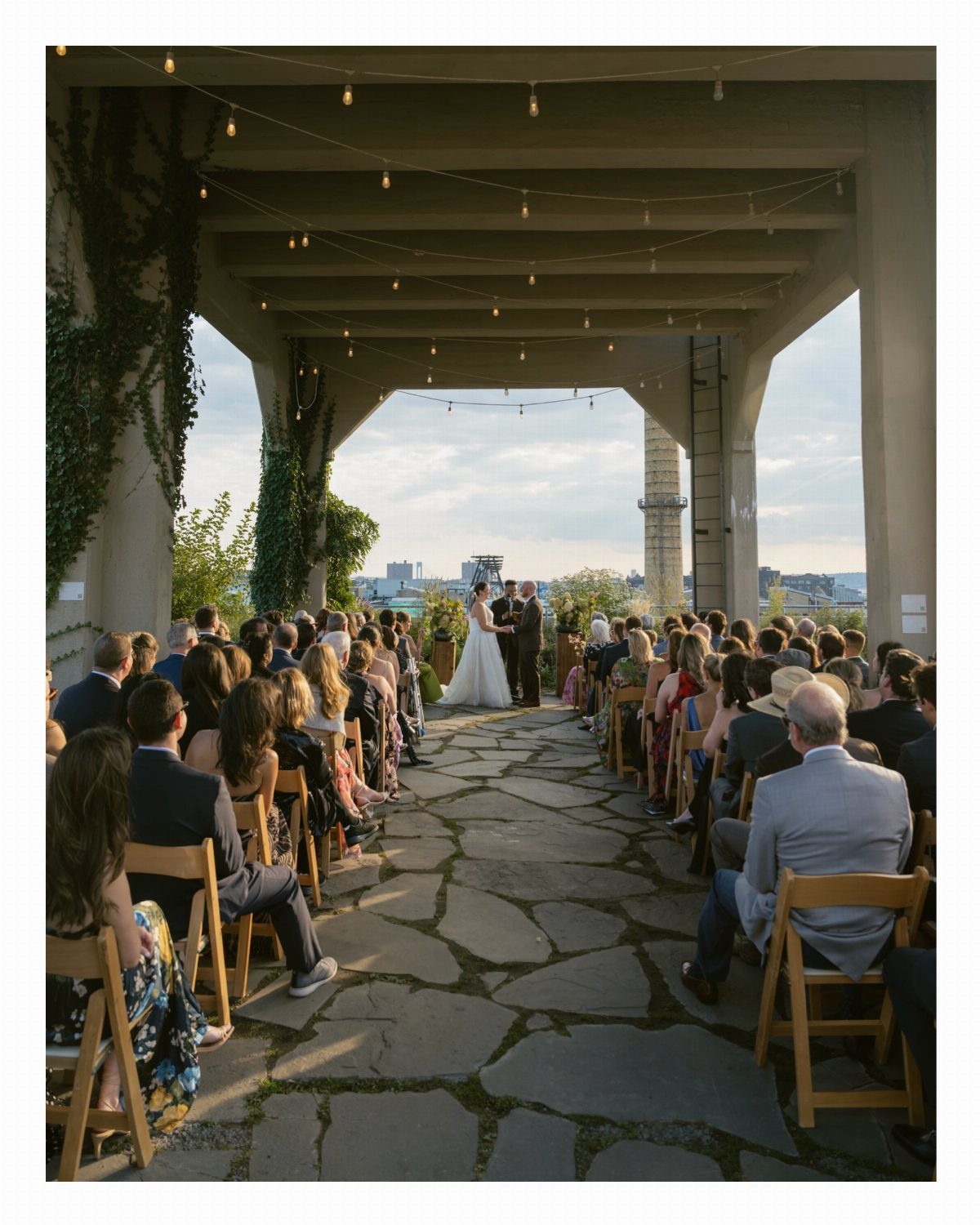 Wedding ceremony under a concrete pergola with guests seated on a stone patio.