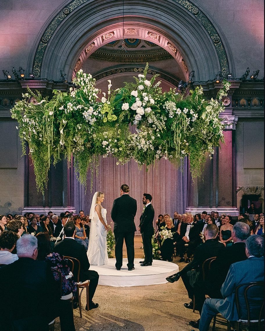Wedding ceremony in a historic hall with a large floating floral installation above the couple.