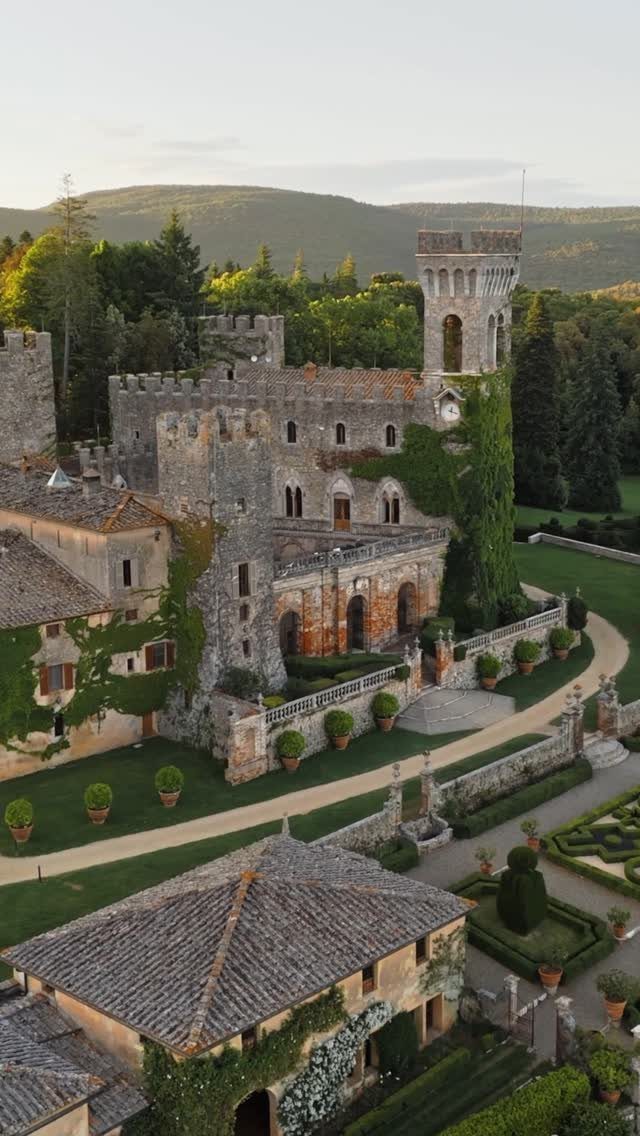 Medieval stone castle with a tall tower and manicured gardens against forested hills.