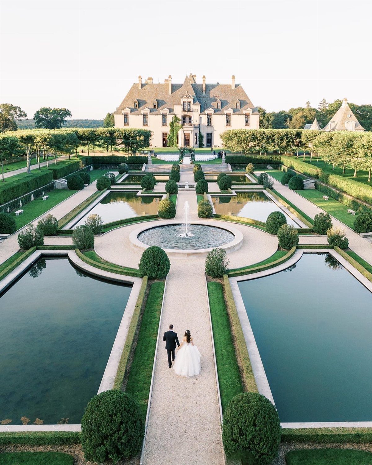 Bride and groom walking toward a grand stone chateau through formal garden pools.