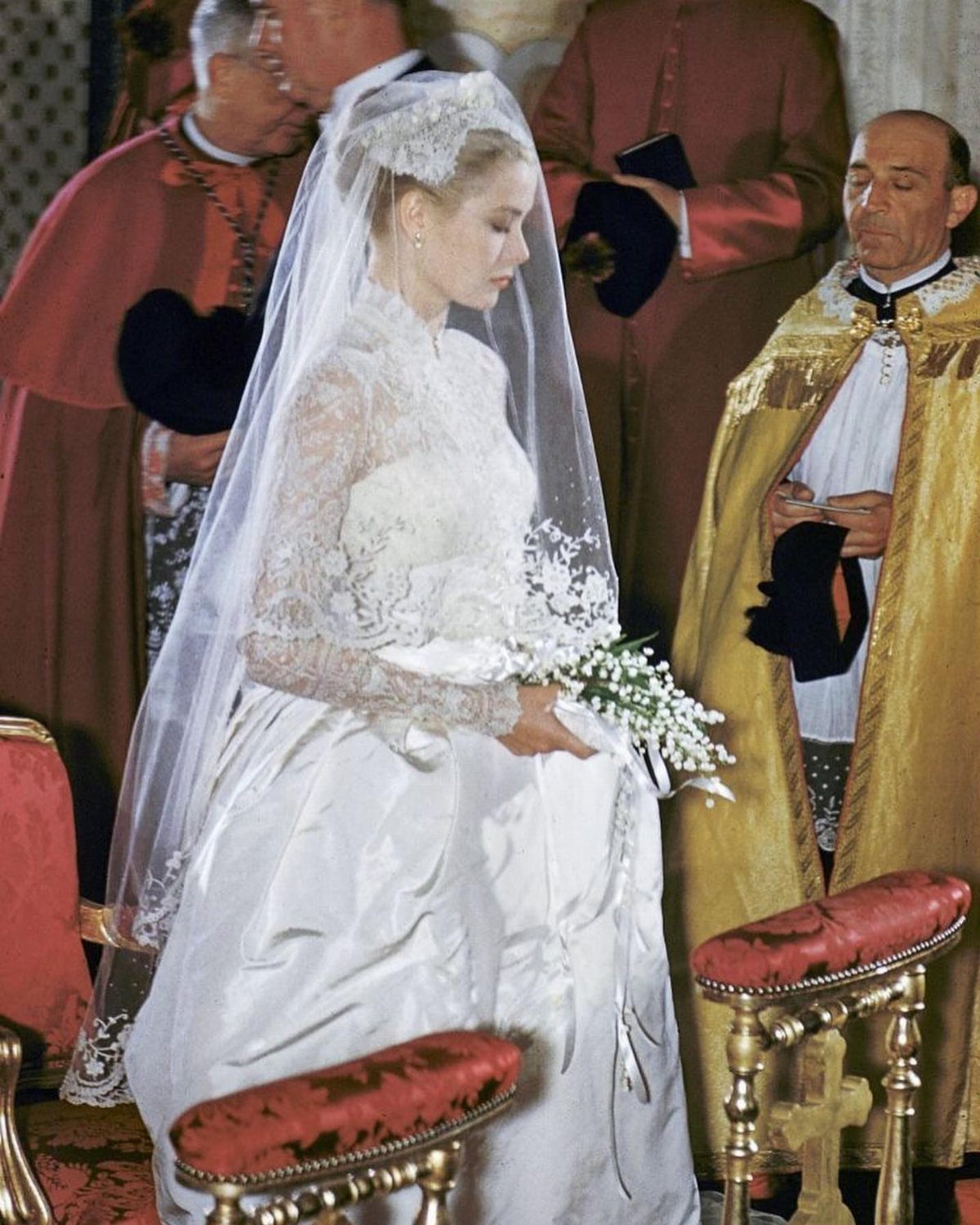 Grace Kelly in her high-necked lace royal wedding dress and veil holding white flowers.