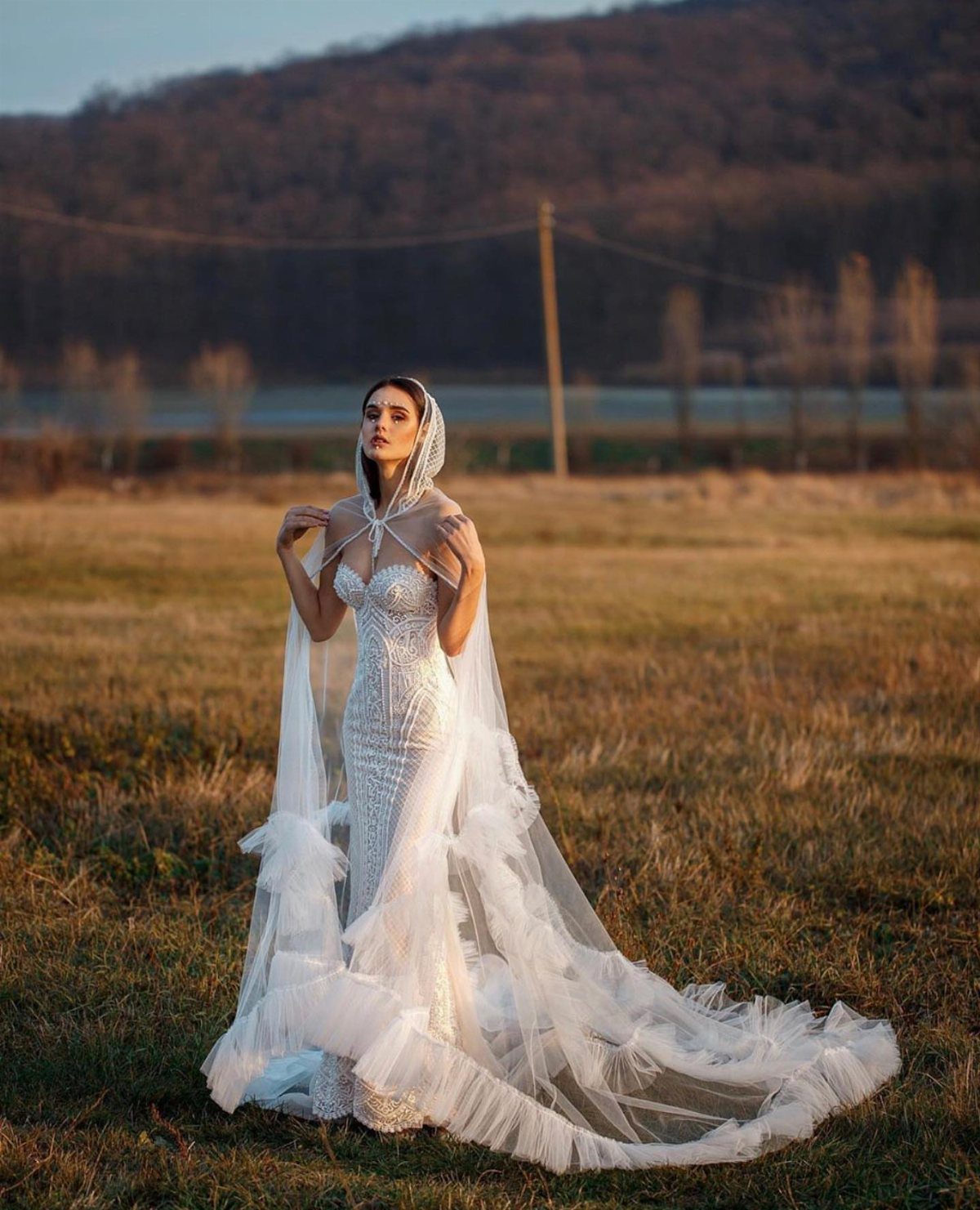 Woman in a white lace wedding gown with a hooded cape in a field