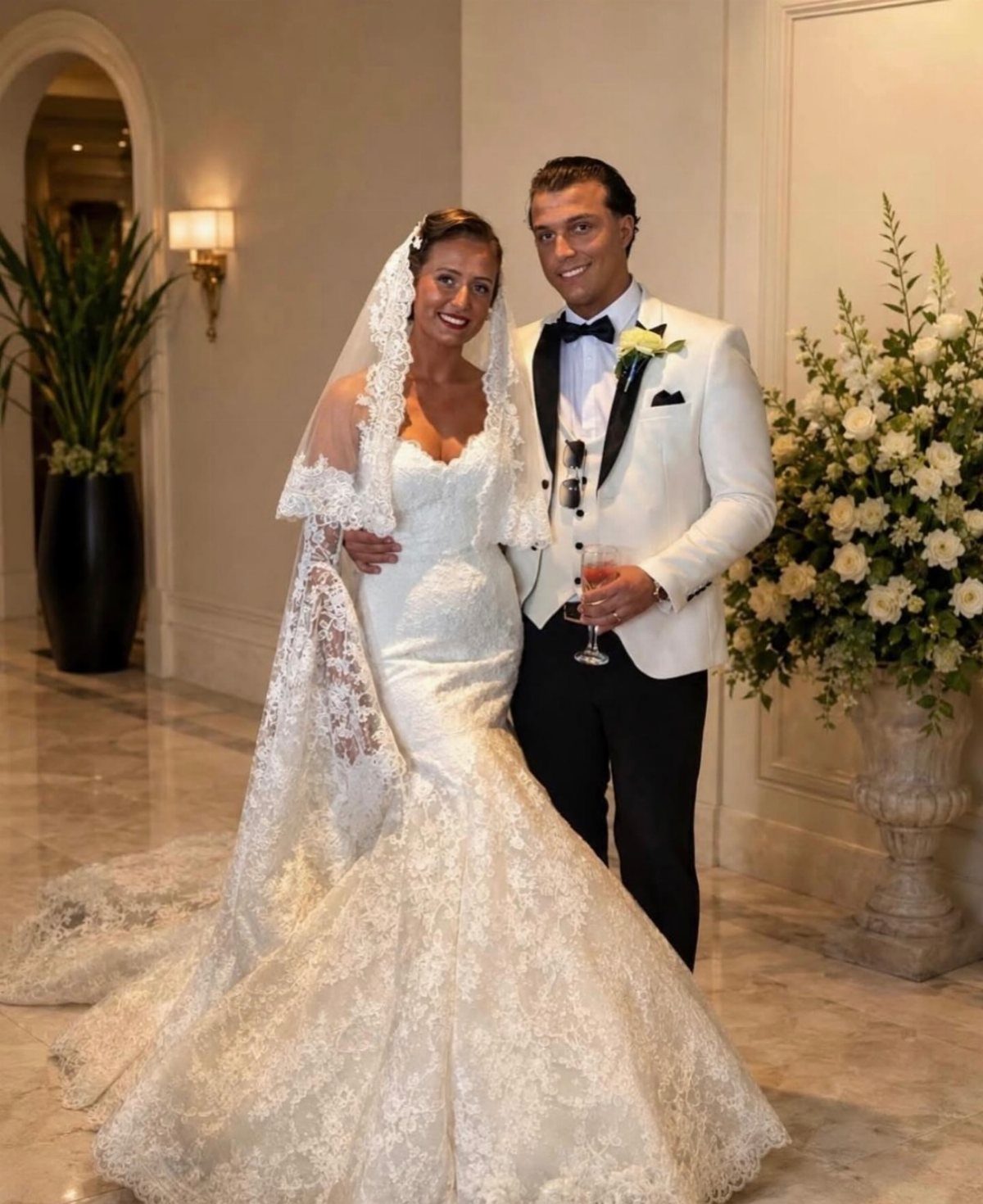 Bride in lace mermaid wedding dress and groom in white tuxedo posing together.