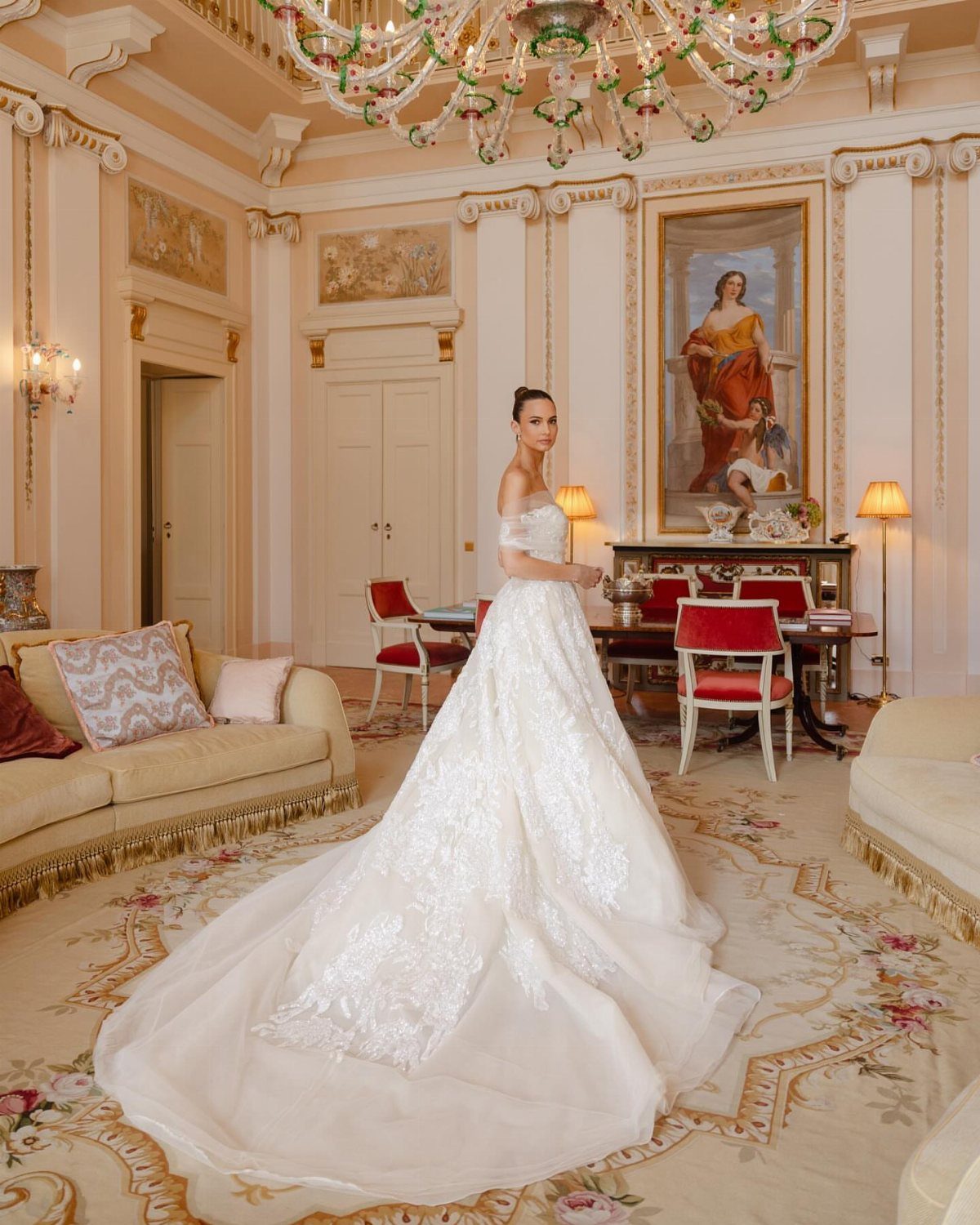 Woman in an off-the-shoulder lace wedding gown standing in an ornate classical room.