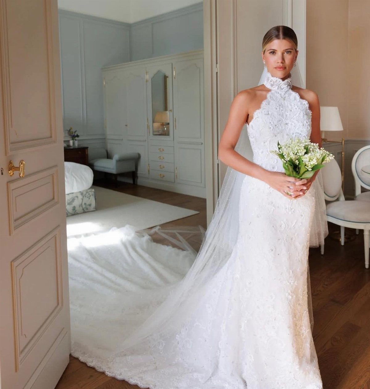 Woman in a high-neck white lace wedding gown holding a small white bouquet.