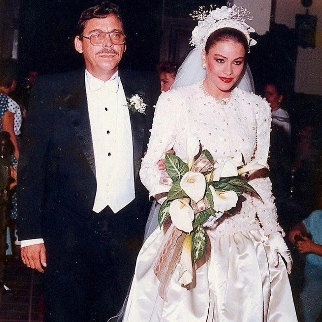 Sofia Vergara in a beaded white wedding gown holding calla lilies next to groom.