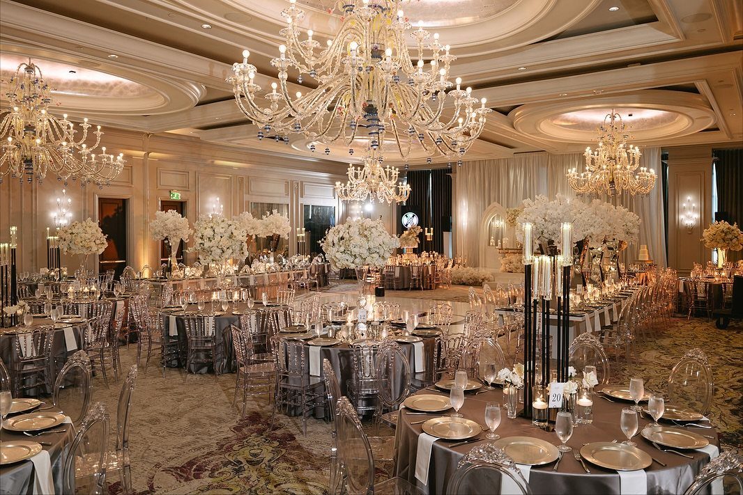 Elegant ballroom with round tables, clear chairs, white flowers, and large crystal chandeliers.