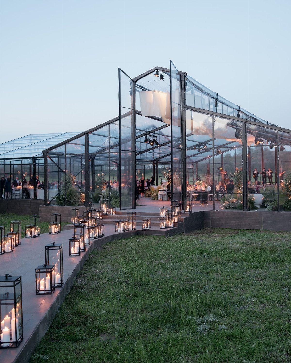 Glass greenhouse event venue with candle lanterns lining a wooden walkway at dusk.