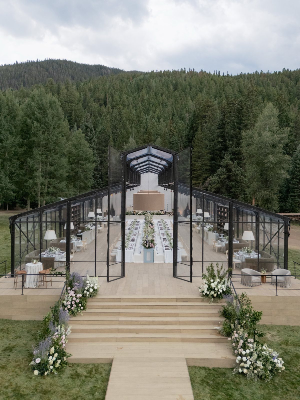 Glass-walled wedding pavilion with dining tables set against a backdrop of forested mountains.