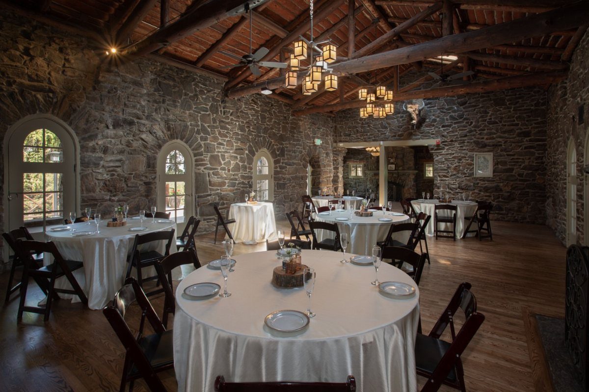 Stone walled event room with round white clothed tables and wooden ceiling beams.