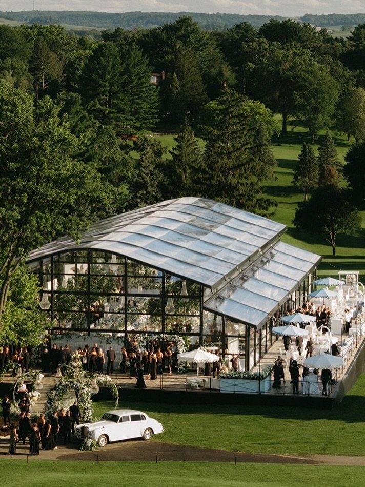 Glass greenhouse wedding venue surrounded by lush green forests and guests in formal wear.