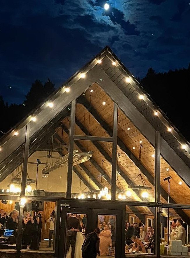 Modern A-frame event venue with glass walls and string lights at night.
