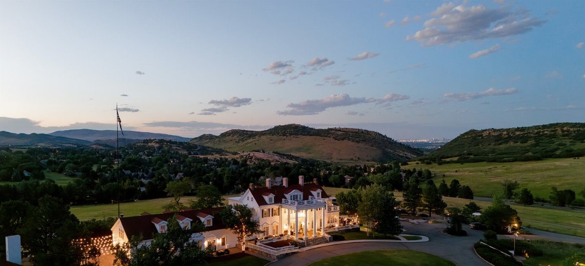Large white colonial estate with a porch set against rolling green hills and mountains.