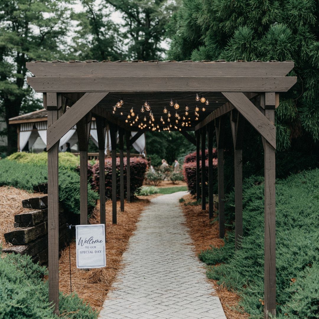 Stone walkway under a wooden pergola with string lights leading to a wedding tent