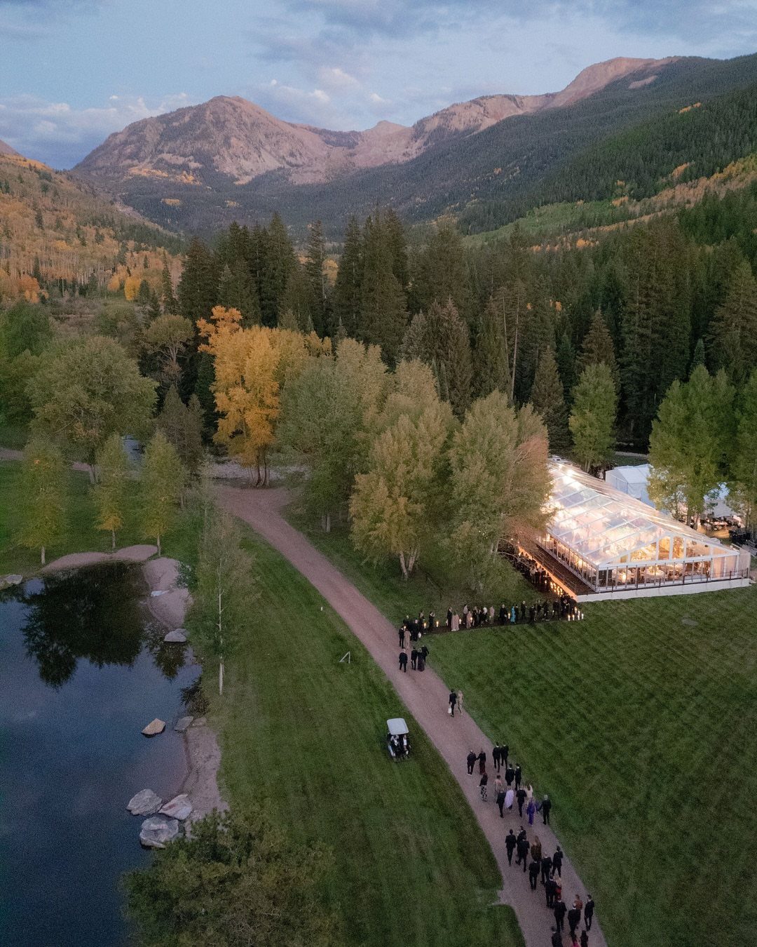 Wedding marquee tent and guests in a mountain valley with autumn trees and pond.