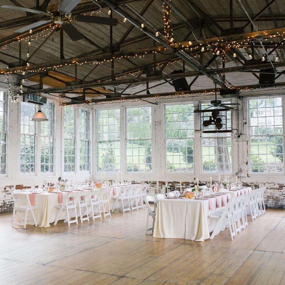 Industrial event space with white dining tables, exposed beams, and large windows.