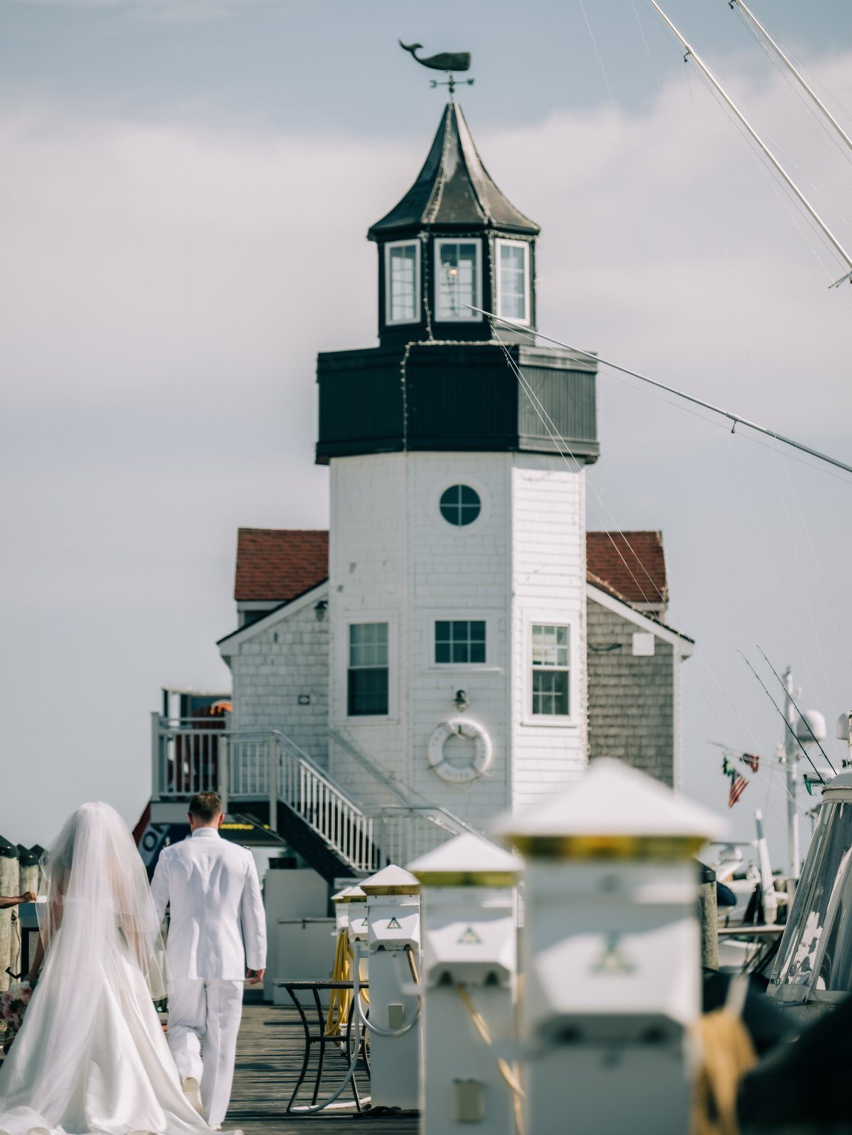 Bride and groom walking on a pier toward a white coastal lighthouse building.