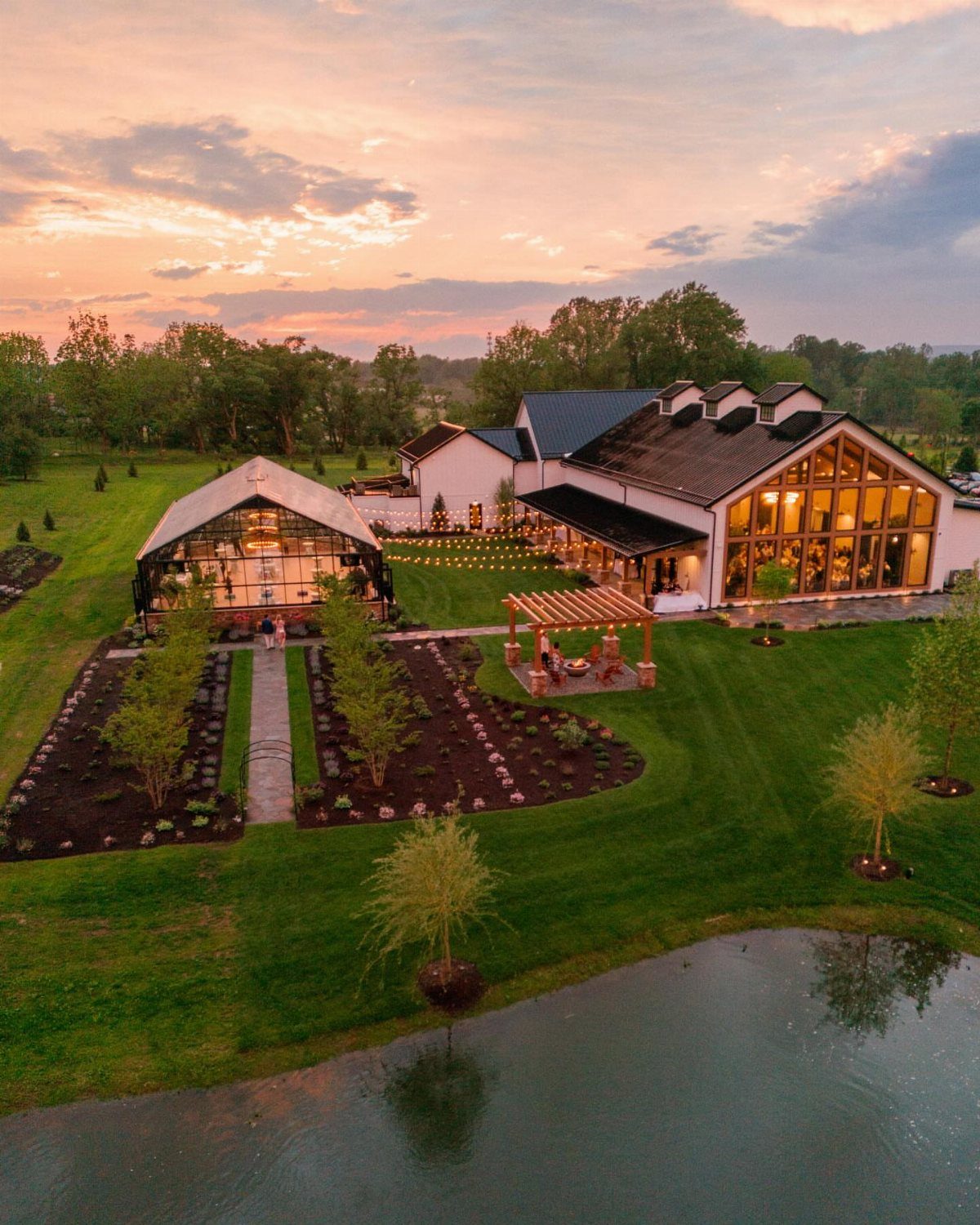 Luxury farm estate with white barns and manicured gardens at sunset by a pond.