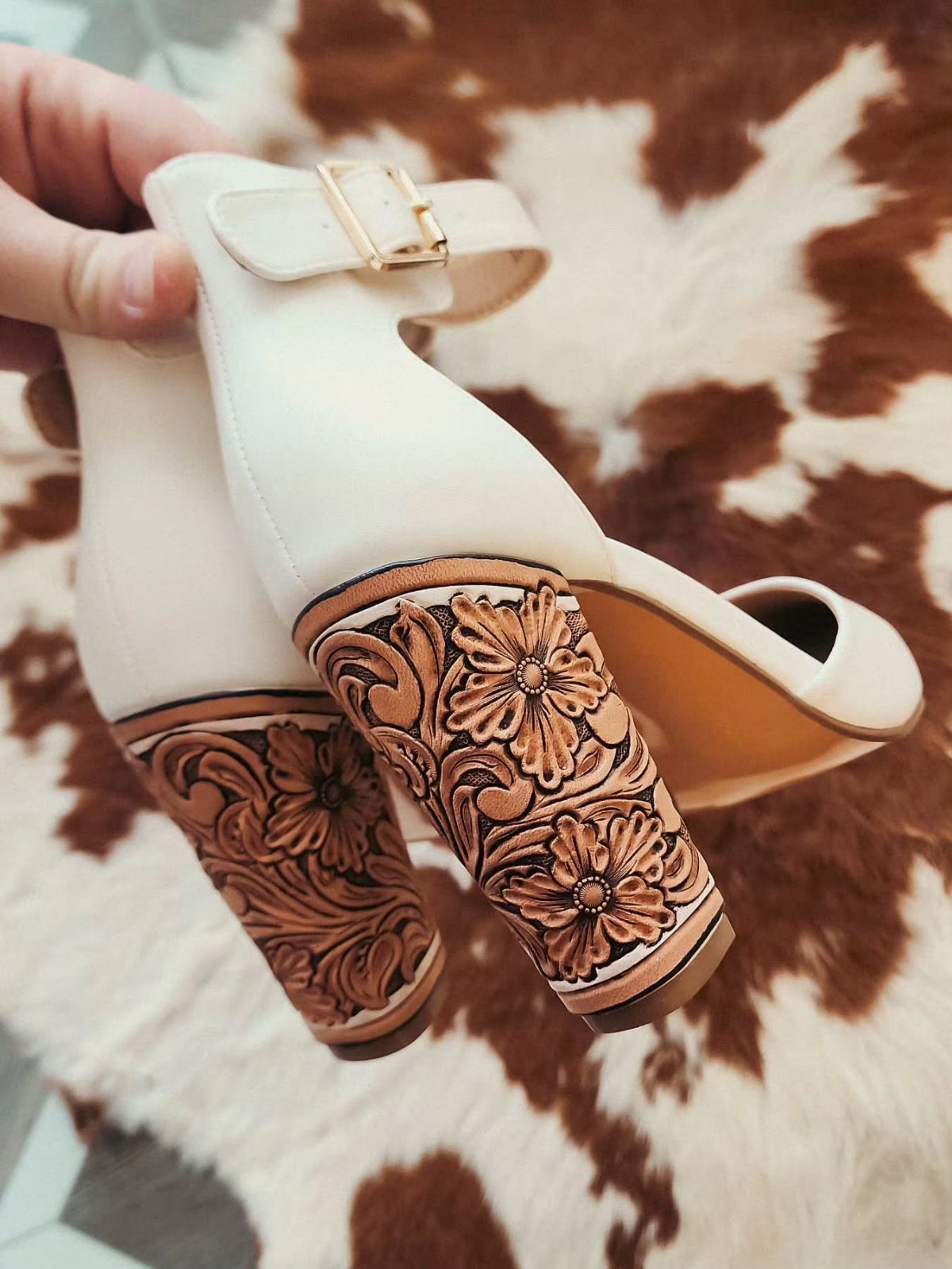 Cream block heels with brown floral tooled leather accents on a cowhide rug.