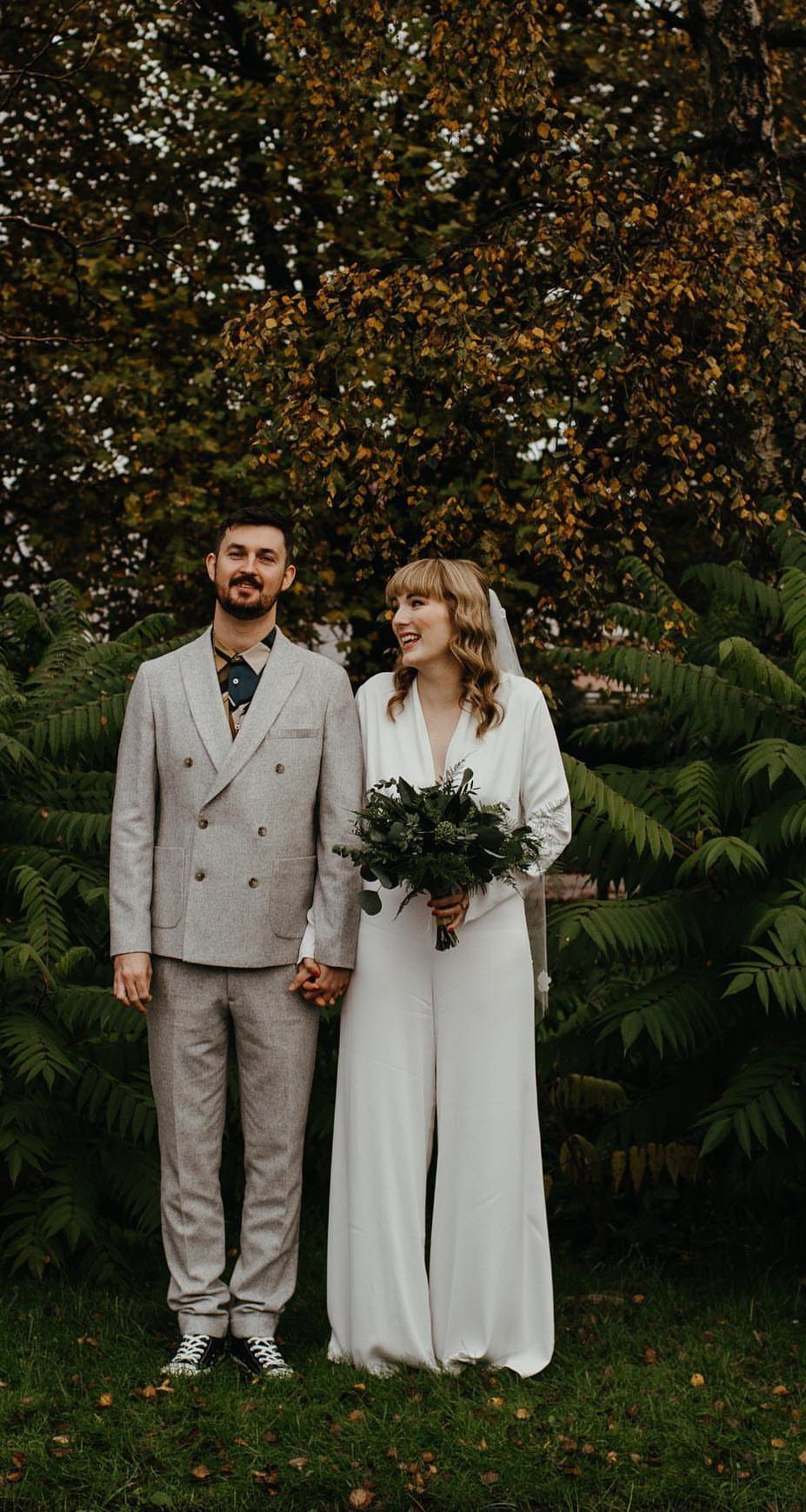 Couple at a wedding, woman in a white dressy jumpsuit holding a bouquet.