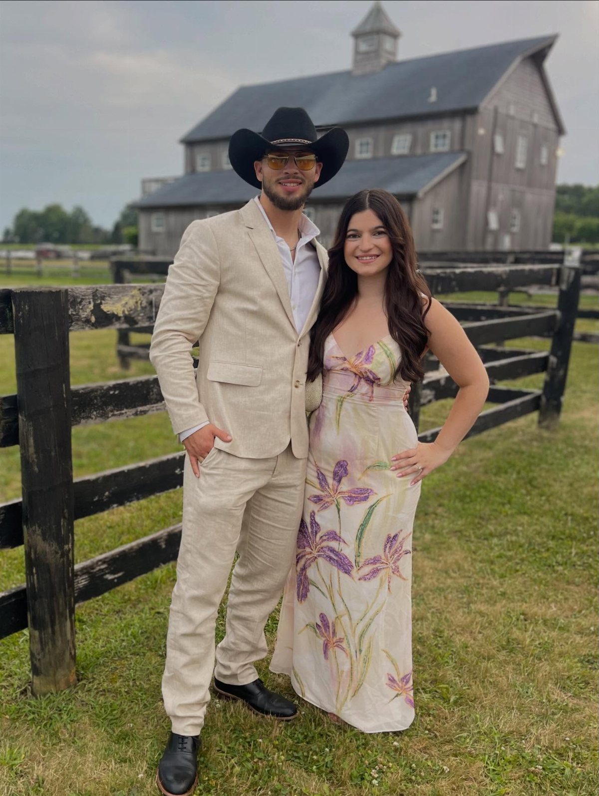 Couple in Western formal attire posing before a large grey barn and fence.