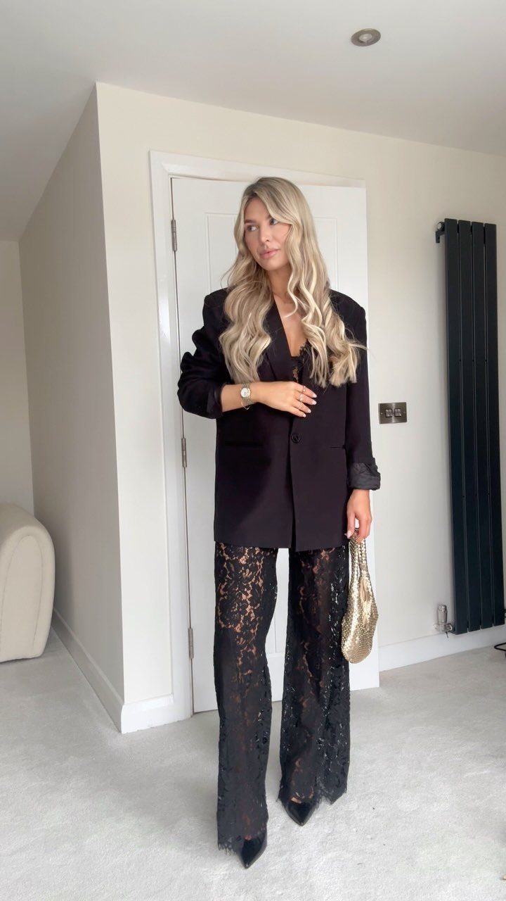 Woman wearing a black blazer and wide-leg lace trousers with a gold clutch.