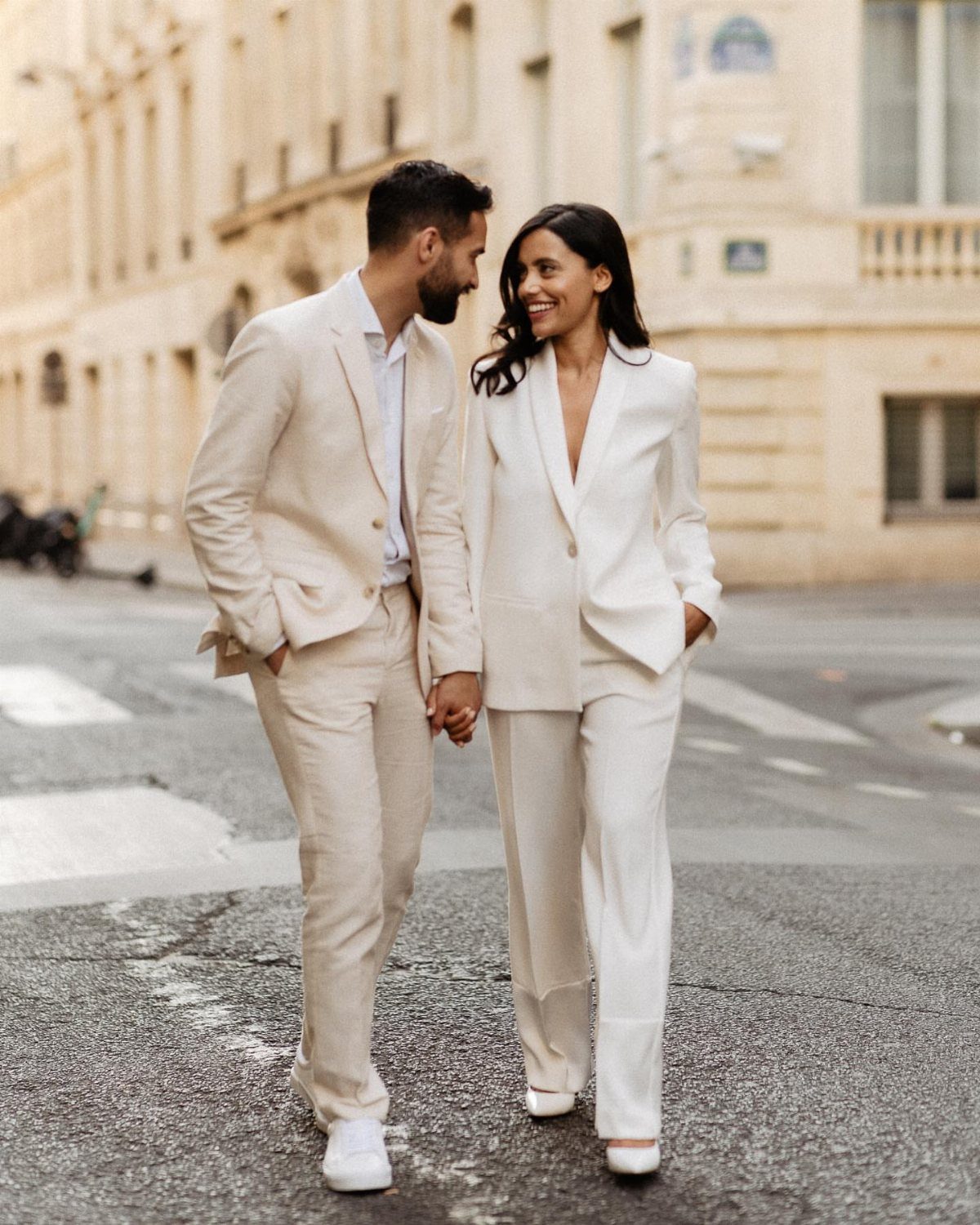 Couple in white and cream suits walking hand-in-hand on a city street.