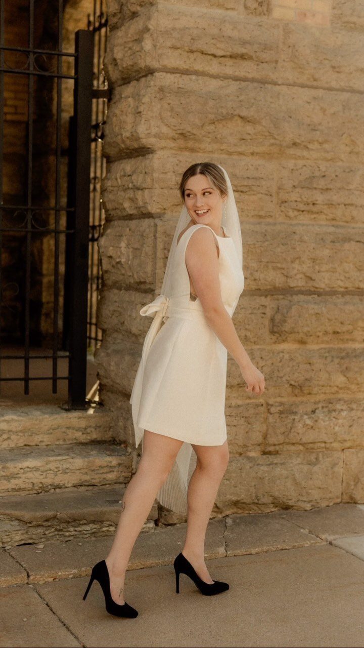Woman in a short white wedding dress and veil posing by a stone wall.